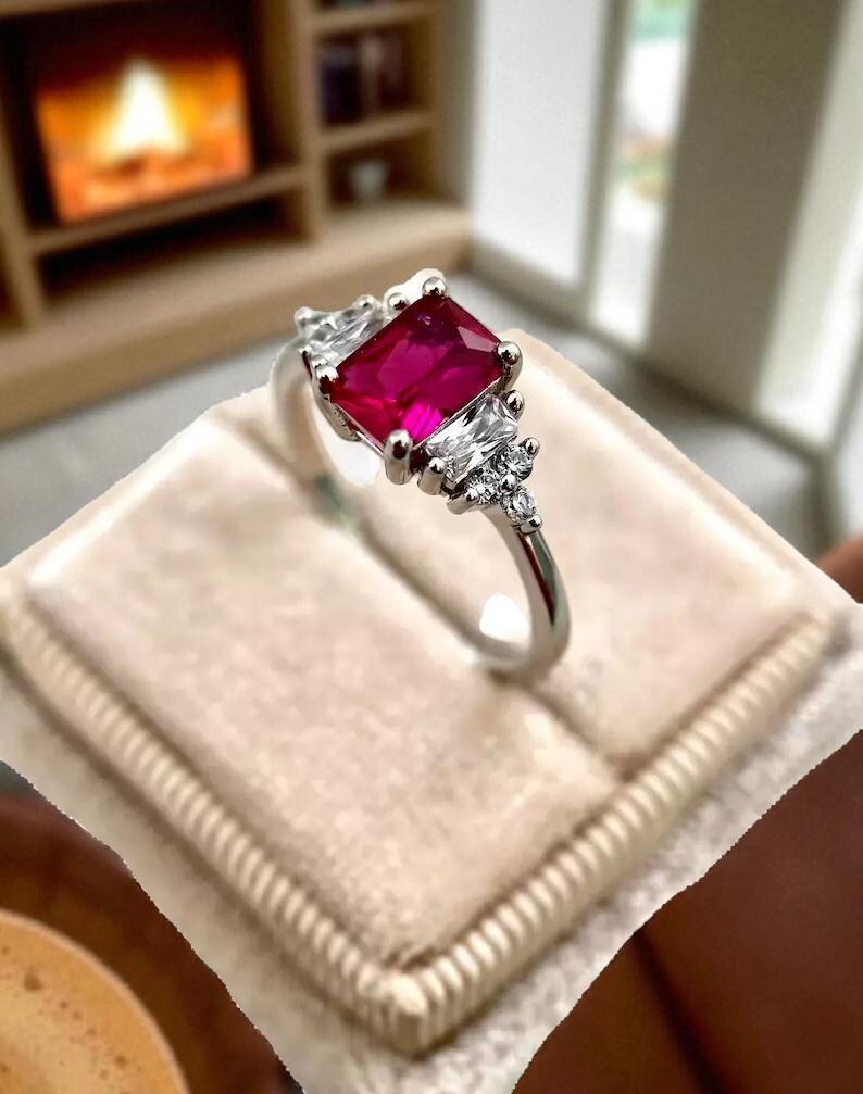 Puede incluir: Un anillo plateado con una gran piedra central de color rub&iacute; cuadrado. El anillo est&aacute; flanqueado por piedras m&aacute;s peque&ntilde;as y transparentes a cada lado y est&aacute; sobre una superficie de terciopelo beige.