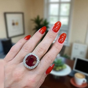 May include: A large, ornate ring featuring a vibrant, oval-cut ruby gemstone, surrounded by a halo of smaller, sparkling diamonds. The ring is set in a silver or white gold band. The nails are painted a bright red.
