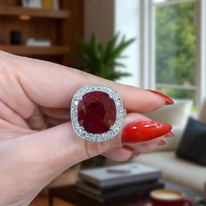 May include: A large, square-cut ruby ring with a halo of small, clear gemstones. The ruby is a deep red color, and the ring's setting is silver-toned. The ring is held up in front of a blurred background.
