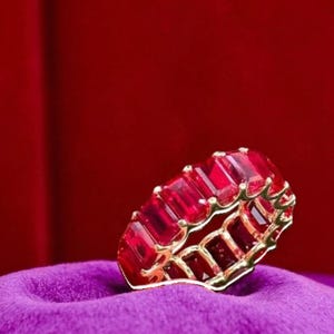 May include: A gold ring with a continuous band of rectangular, deep red gemstones. The ring is set against a vibrant purple fabric and a rich red background, highlighting the ring's color and design.