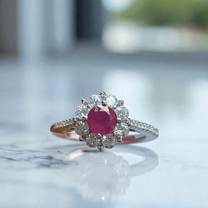 May include: A silver ring with a central, round, deep red gemstone surrounded by a halo of smaller, clear gemstones. The ring's band is also adorned with small, clear gemstones, creating a sparkling effect. The ring is set against a blurred background.