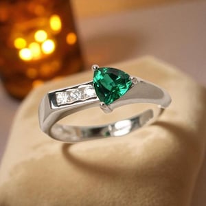 Op de afbeelding: Een zilveren ring met een driehoekige, smaragdgroene edelsteen. De band heeft een rechthoekig ontwerp met drie kleinere, heldere edelstenen. De ring is geplaatst tegen een zachte, neutrale achtergrond.
