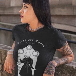 May include: Black t-shirt with a white graphic design featuring a woman in a hooded cloak and the text "Liars are Fools, What is false?" and "Skepticism."
