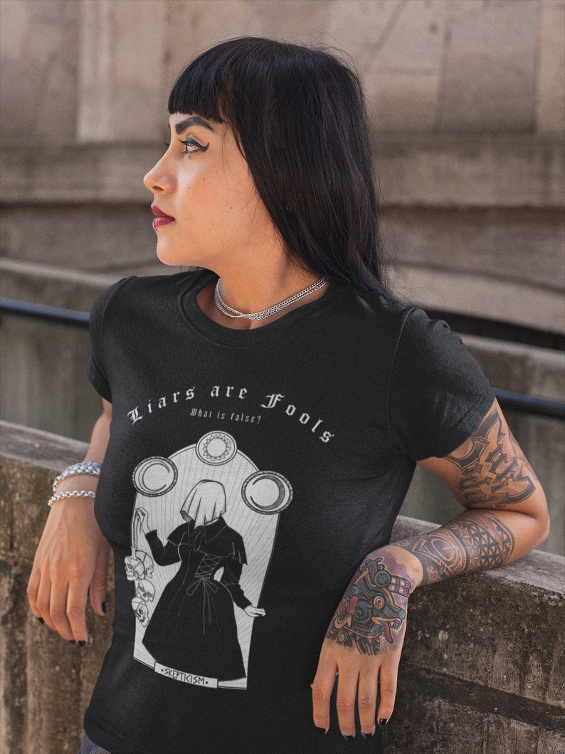 May include: Black t-shirt with a white graphic design featuring a woman in a hooded cloak and the text "Liars are Fools, What is false?" and "Skepticism."