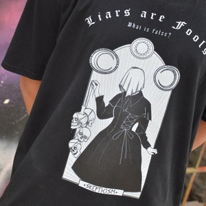 May include: Black t-shirt with a white graphic print of a woman in a hooded dress holding a chain of skulls. The text "Liars are Fools" is printed above the graphic and "What is false?" is printed below the graphic. The text "Skepticism" is printed below the graphic.
