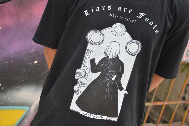 May include: Black t-shirt with a white graphic print of a woman in a hooded dress holding a chain of skulls. The text "Liars are Fools" is printed above the graphic and "What is false?" is printed below the graphic. The text "Skepticism" is printed below the graphic.