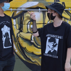 May include: Two people wearing black t-shirts with graphic designs. The person on the left is wearing a black bucket hat and a blue face mask. The person on the right is wearing a black bucket hat and a black face mask. The t-shirt on the left has a black and white illustration of a person in a long dress with the text "Lights are low" and "Be careful". The t-shirt on the right has a black and white illustration of a person with the text "Antisocial Vampire Club".