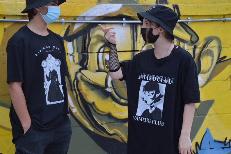 May include: Two people wearing black t-shirts with graphic designs. The person on the left is wearing a black bucket hat and a blue face mask. The person on the right is wearing a black bucket hat and a black face mask. The t-shirt on the left has a black and white illustration of a person in a long dress with the text "Lights are low" and "Be careful". The t-shirt on the right has a black and white illustration of a person with the text "Antisocial Vampire Club".