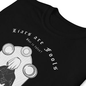 May include: Black t-shirt with a white graphic design. The graphic features a hooded figure with a crescent moon and sun symbols. The text "Liars are Fools" is written in a gothic font above the figure. The text "What is false?" is written below the figure.