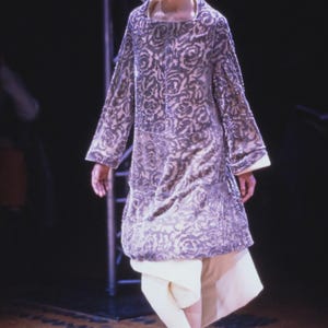 Puede incluir: Un vestido largo de manga larga, hasta la rodilla, con un estampado floral en tonos grises y morados. El vestido se combina con pantalones color crema y zapatos negros. La modelo est&aacute; sobre un suelo de madera.