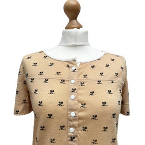 May include: A short-sleeved, beige button-down shirt with a black repeating pattern. The shirt has a white button placket.