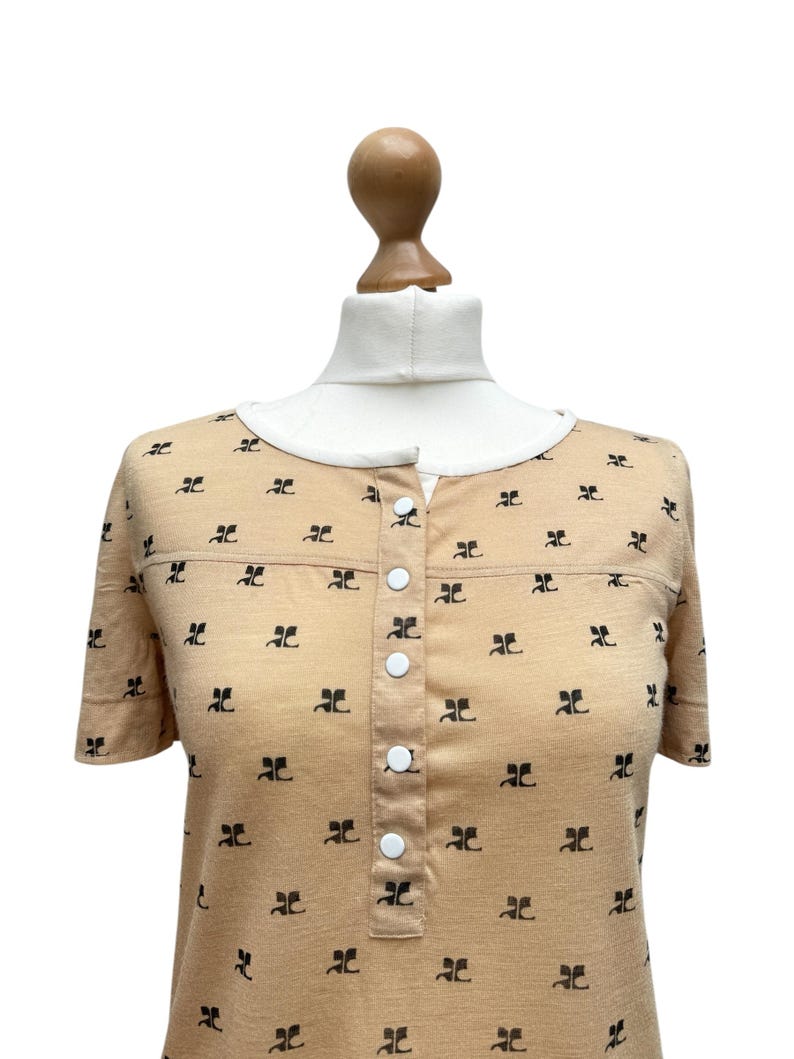 May include: A short-sleeved, beige button-down shirt with a black repeating pattern. The shirt has a white button placket.