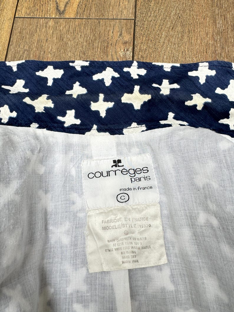 K&ouml;nnte beinhalten: Nahaufnahme des Inneren eines Kleidungsst&uuml;cks mit marineblauem Stoff und einem wei&szlig;en Kreuzmuster. Das Futter ist wei&szlig;. Ein Etikett lautet "courreges paris made in france" und enth&auml;lt Waschanweisungen.