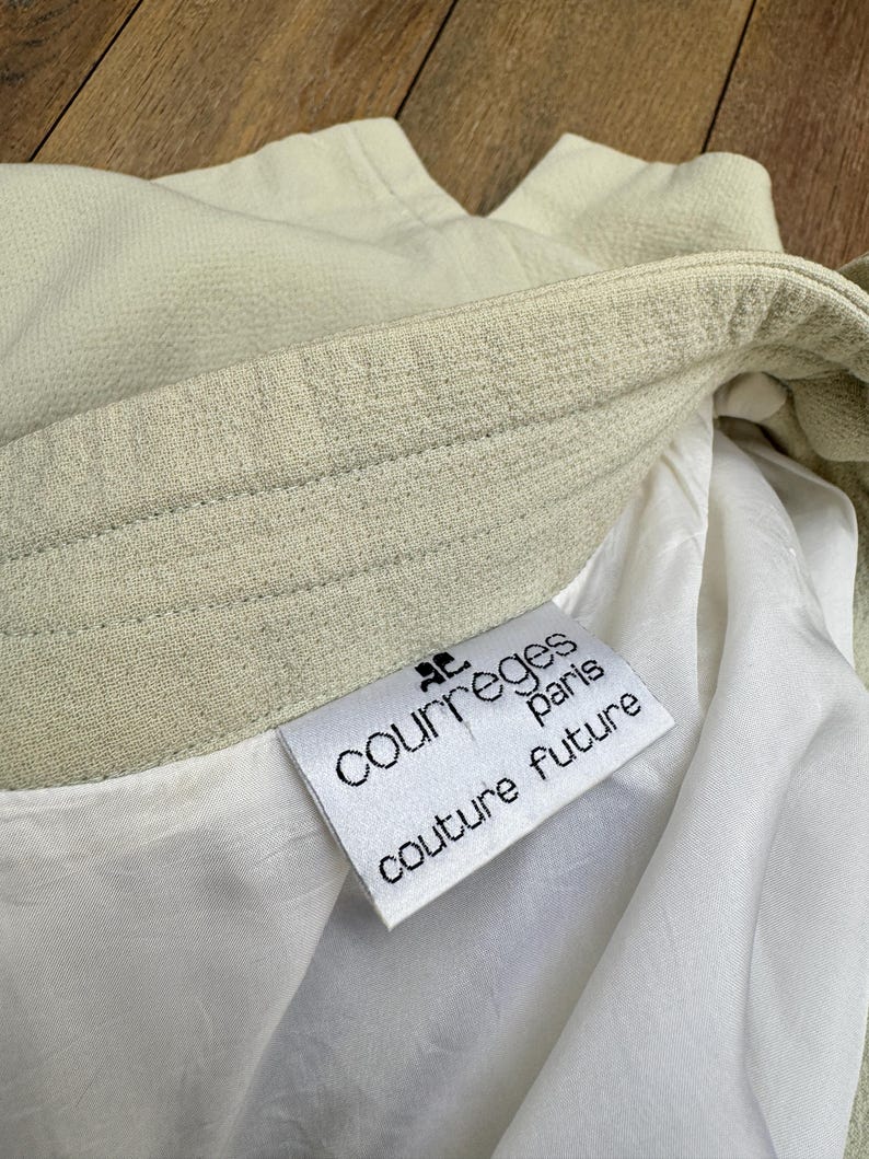 Puede incluir: Primer plano de una prenda verde claro con forro blanco. La etiqueta dice "COURREGES paris couture future" en texto negro. La tela tiene una apariencia texturizada.