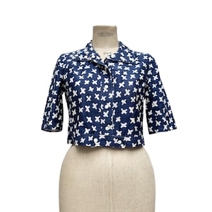 K&ouml;nnte beinhalten: Eine marineblaue, kurze Jacke mit wei&szlig;em Kreuzmuster. Die Jacke hat kurze &Auml;rmel, einen Kragen und Kn&ouml;pfe vorne. Das Kleidungsst&uuml;ck wird auf einer Vintage-Schneiderpuppe pr&auml;sentiert.