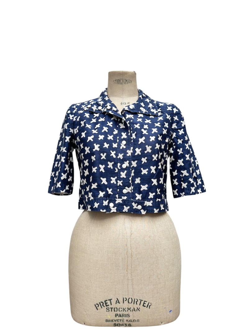 K&ouml;nnte beinhalten: Eine marineblaue, kurze Jacke mit wei&szlig;em Kreuzmuster. Die Jacke hat kurze &Auml;rmel, einen Kragen und Kn&ouml;pfe vorne. Das Kleidungsst&uuml;ck wird auf einer Vintage-Schneiderpuppe pr&auml;sentiert.