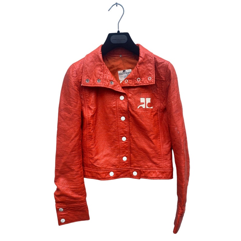 May include: A bright orange leather jacket with a white logo on the front. The jacket has a snap closure and a collar that can be folded up.