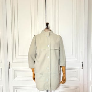 Puede incluir: Un abrigo vintage beige claro con cuello alto y cierre de botones. El abrigo tiene mangas tres cuartos y dos bolsillos delanteros. Se exhibe en un maniqu&iacute; de madera.