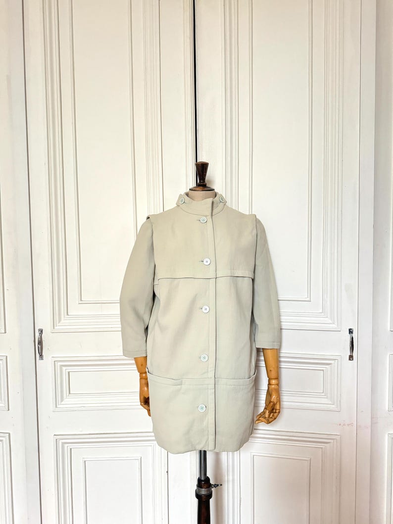 Puede incluir: Un abrigo vintage beige claro con cuello alto y cierre de botones. El abrigo tiene mangas tres cuartos y dos bolsillos delanteros. Se exhibe en un maniqu&iacute; de madera.
