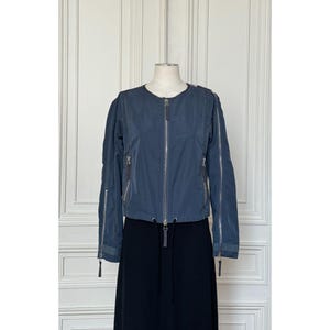 May include: A slate blue zip-up jacket featuring a round neckline, long sleeves with full-length vertical zippers, and two front zippered pockets. The jacket has a drawstring hem with silver-toned hardware and a central front zipper closure.