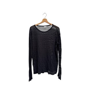 May include: Black long-sleeved mesh shirt hanging on a wooden hanger. The shirt has a round neckline and a loose, see-through weave. The sleeves are long and the fabric appears lightweight.