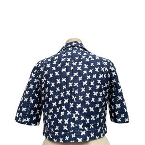 K&ouml;nnte beinhalten: Marineblaue Jacke mit wei&szlig;en Kreuzmustern. Die Jacke hat kurze &Auml;rmel und einen Kragen. Sie wird auf einer Schaufensterpuppe pr&auml;sentiert, um das Design und die Passform hervorzuheben. Der Stoff scheint strukturiert zu sein.