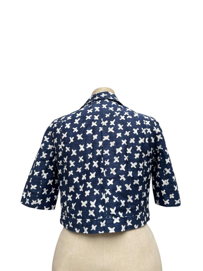 K&ouml;nnte beinhalten: Marineblaue Jacke mit wei&szlig;en Kreuzmustern. Die Jacke hat kurze &Auml;rmel und einen Kragen. Sie wird auf einer Schaufensterpuppe pr&auml;sentiert, um das Design und die Passform hervorzuheben. Der Stoff scheint strukturiert zu sein.