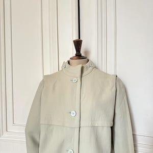 Puede incluir: Un abrigo tipo trench de color beige claro con cuello alto y cierre de botones. El abrigo presenta un detalle &uacute;nico de capa en los hombros y botones azul claro. El abrigo se exhibe en un maniqu&iacute; de madera.