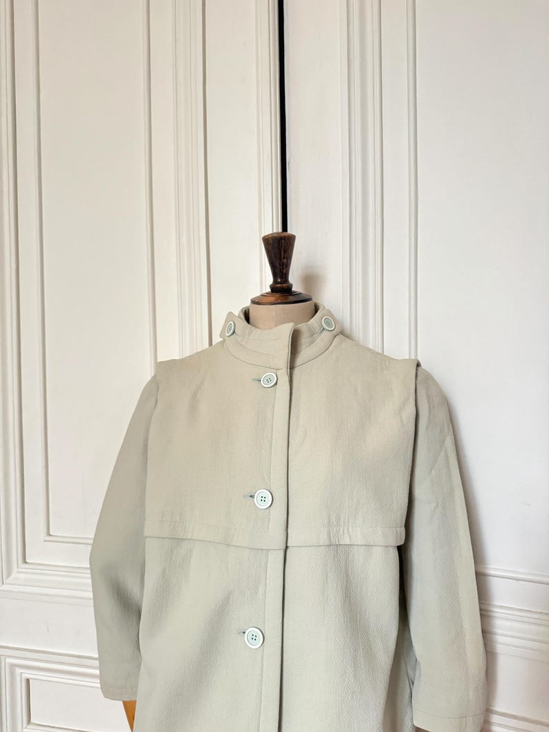 Puede incluir: Un abrigo tipo trench de color beige claro con cuello alto y cierre de botones. El abrigo presenta un detalle &uacute;nico de capa en los hombros y botones azul claro. El abrigo se exhibe en un maniqu&iacute; de madera.