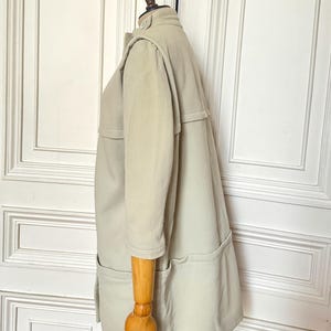 Puede incluir: Un abrigo tipo gabardina de color beige claro con mangas tres cuartos, cuello abotonado y dos bolsillos delanteros. El abrigo se exhibe en un maniqu&iacute; de madera, mostrando su dise&ntilde;o a medida y color neutro.