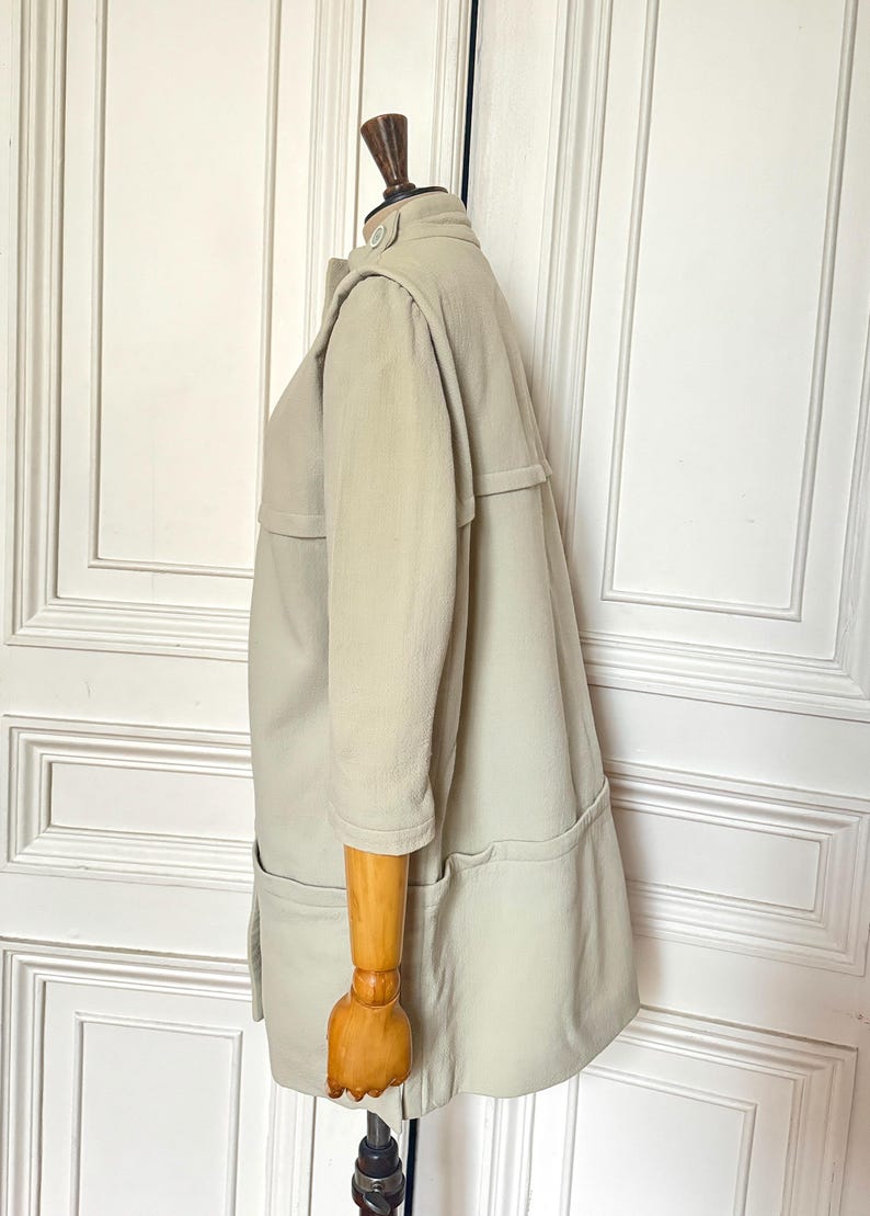 Puede incluir: Un abrigo tipo gabardina de color beige claro con mangas tres cuartos, cuello abotonado y dos bolsillos delanteros. El abrigo se exhibe en un maniqu&iacute; de madera, mostrando su dise&ntilde;o a medida y color neutro.