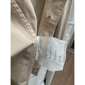 May include: A beige cotton jacket with a white label that reads "100% Baumwolle 100% Algodon 100% Cotton" and "See reverse for care instruction".