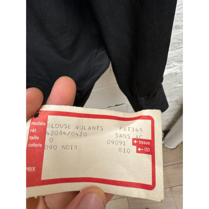 May include: A white tag with red trim and black text. The tag is labeled "BLOUSE VOLANTS 42084/0420" and "PAT345 SANS AC 09091 810" with the words "tissus" and "OD" written in red.