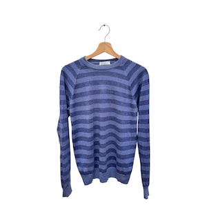 Peut inclure: Un pull bleu à rayures avec des manches longues est suspendu à un cintre en bois. Le pull présente des rayures horizontales dans différentes nuances de bleu. Le vêtement est fait d'une matière douce et légère, adaptée au superposition ou à porter seul.