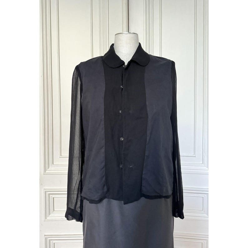 Pode incluir: Camisa preta abotoada com gola redonda. A camisa apresenta mangas compridas e transparentes nas laterais e um painel central liso. A pe&ccedil;a &eacute; exibida em um manequim, mostrando seu ajuste e design sob medida.