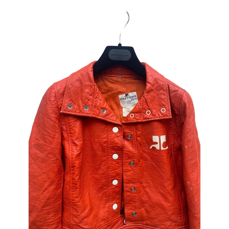 May include: A bright orange leather jacket with a white logo that says "Courr&egrave;ges Paris". The jacket has a high collar and a button closure.