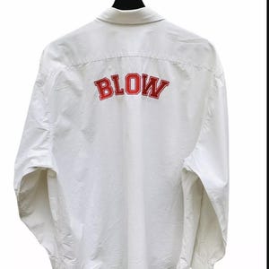 May include: White button-down shirt with the word "BLOW" in red, collegiate-style lettering across the back. The shirt is hanging on a black hanger against a white background. The shirt has long sleeves and a collar.