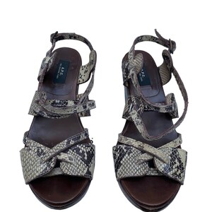 May include: A pair of brown and beige snake-print sandals. The sandals have multiple straps, a buckle closure, and a dark brown sole. The brand name "A.P.C." is visible on the inner sole. The shoes are set against a white background.