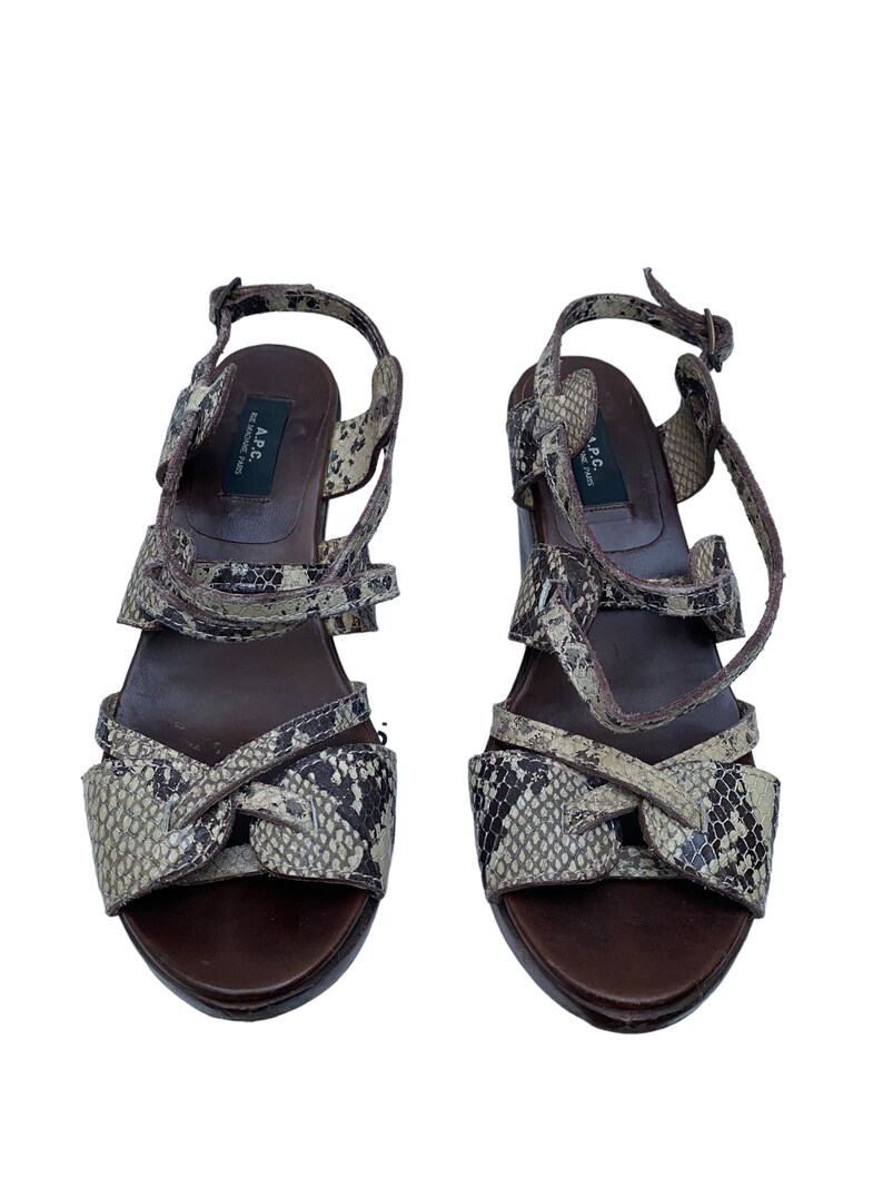 May include: A pair of brown and beige snake-print sandals. The sandals have multiple straps, a buckle closure, and a dark brown sole. The brand name "A.P.C." is visible on the inner sole. The shoes are set against a white background.