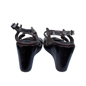 May include: Pair of wedge sandals with a snakeskin pattern on the straps and a dark brown wooden wedge heel. The sandals have an ankle strap with a buckle closure.