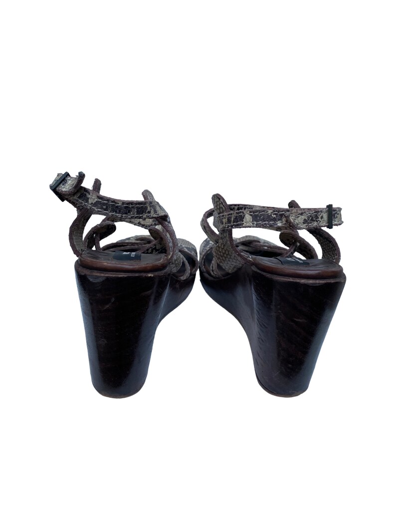 May include: Pair of wedge sandals with a snakeskin pattern on the straps and a dark brown wooden wedge heel. The sandals have an ankle strap with a buckle closure.