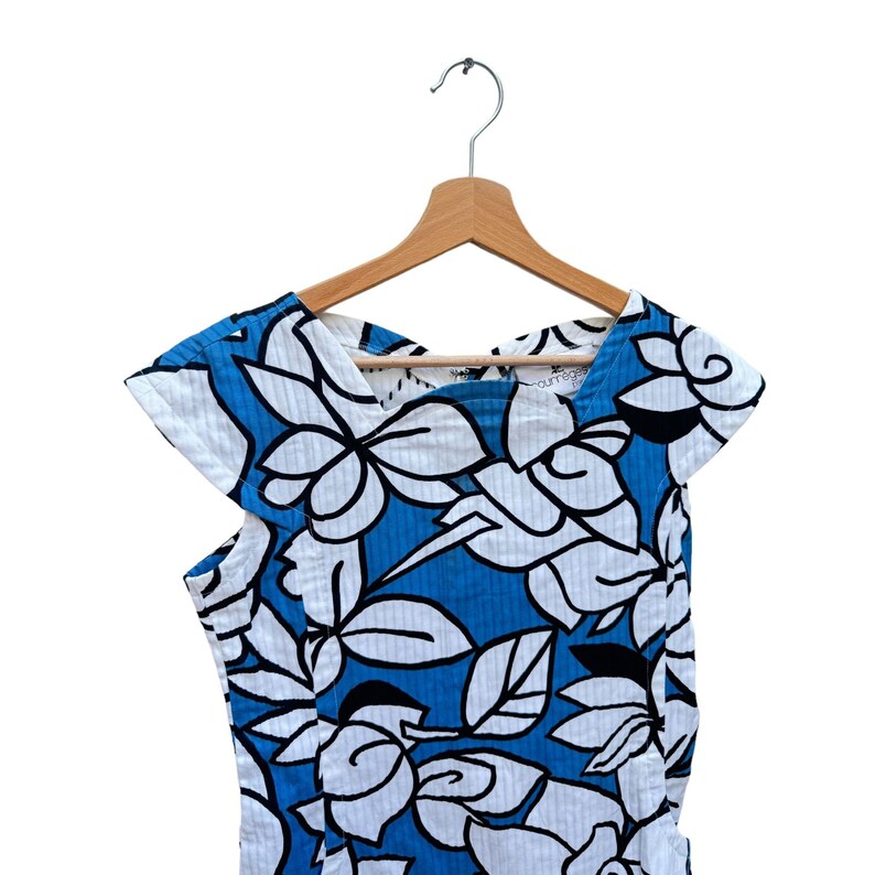 Puede incluir: Un vestido de estampado floral azul y blanco con mangas cortas colgado de una percha de madera. El vestido tiene cuello en V y un corte holgado.