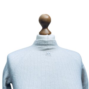 May include: A light blue ribbed turtleneck sweater with a small white button on the front.