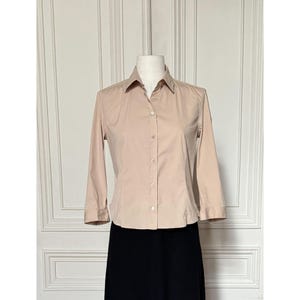 Peut inclure: Une chemise boutonnée beige clair à manches trois-quarts. La chemise est présentée sur un mannequin, associée à une jupe noire. La chemise a un col classique et une fermeture boutonnée.