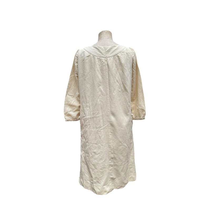 Pode incluir: Um vestido de manga comprida de cor creme com decote redondo e um detalhe franzido na gola. O vestido tem um caimento solto e uma bainha ligeiramente franzido.
