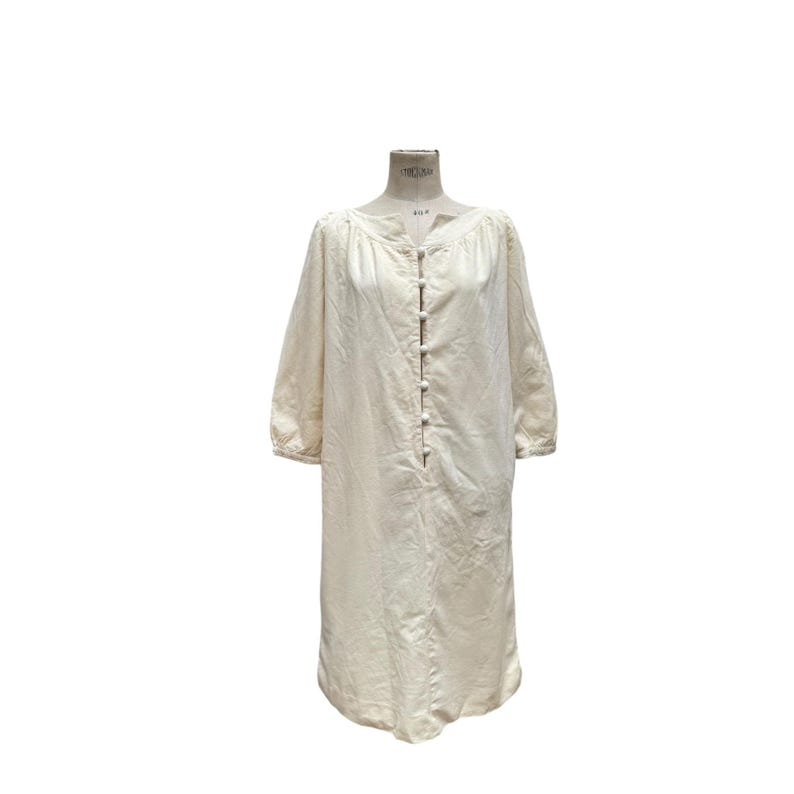 Pode incluir: Um vestido comprido de linho branco-queimado com decote redondo e fecho de bot&otilde;es na frente. O vestido tem um caimento solto e uma cintura ligeiramente franzida.