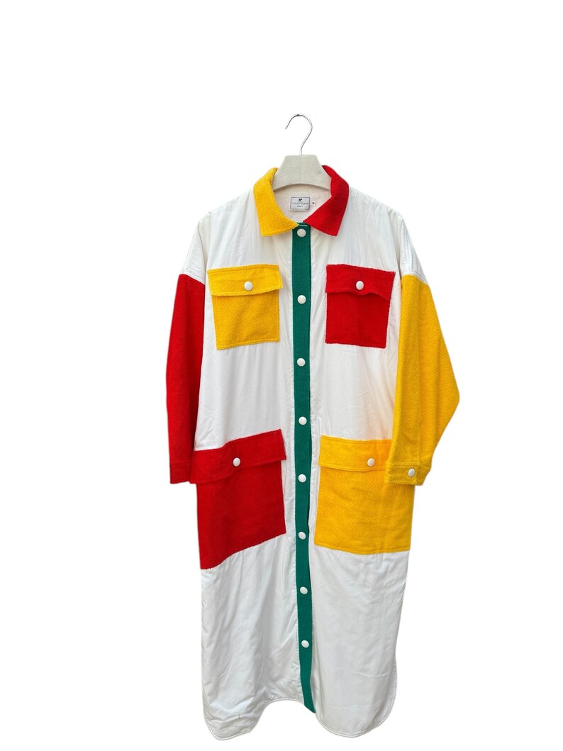 Puede incluir: Una camisa blanca de manga larga con un frente abotonado verde. La camisa tiene grandes cuadrados rojos, amarillos y verdes cosidos en la parte delantera y las mangas.