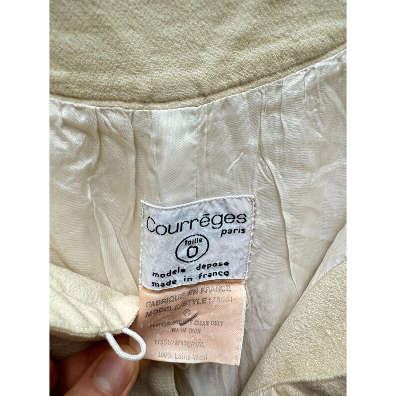 Pode incluir: Um casaco de l&atilde; branco com uma etiqueta que diz "Courr&egrave;ges Paris" e "Taille 0" com as palavras "mod&egrave;le depose made in France" e "Fabriqu&eacute; en France" abaixo. A etiqueta tamb&eacute;m inclui o texto "MODELS/STYLE: 28061", "Genression CLEAN ONLY", "WAIM IRON", "ISSU/MAYERIAL" e "100% Laine Wool".