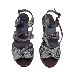 May include: A pair of brown and snakeskin-patterned sandals. The sandals have multiple straps, a bow detail on the front, and a buckle closure. The soles are dark brown.