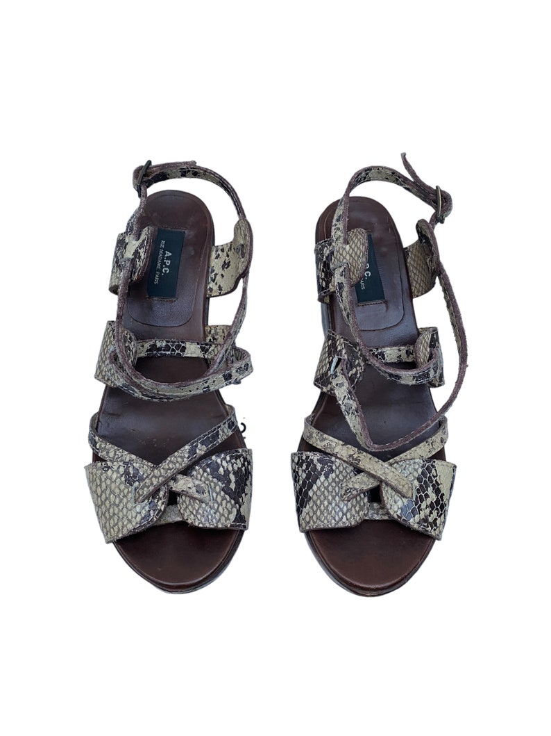May include: A pair of brown and snakeskin-patterned sandals. The sandals have multiple straps, a bow detail on the front, and a buckle closure. The soles are dark brown.